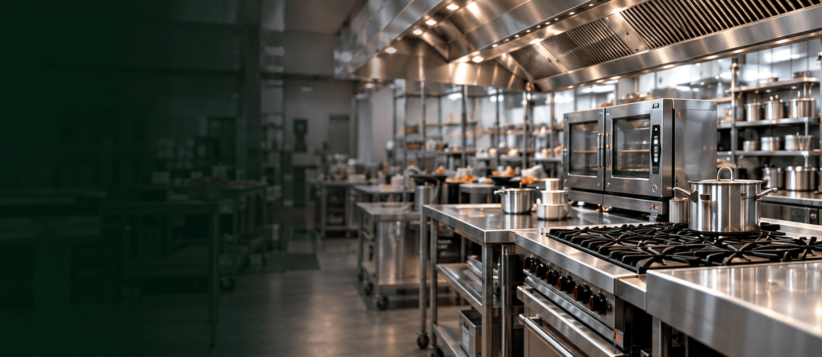 A professional commercial kitchen with stainless steel gas ranges, ovens, workstations, and a large ventilation hood under warm overhead lighting.